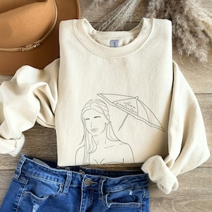 May include: A cream colored sweatshirt with a black line drawing of a woman with long hair under a beach umbrella. The umbrella has the words "Sunscreen Protection" written on it.