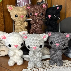 May include: Six crocheted cat plushies in various colors, including beige, brown, black, white, and gray. Each plush has pink accents on the nose and ears.