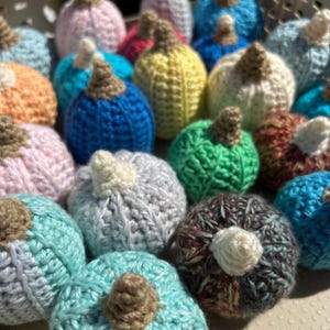 May include: A collection of colorful, handmade crochet pumpkins in various shades including blue, green, pink, and yellow. Each pumpkin has a brown stem, perfect for fall decor.