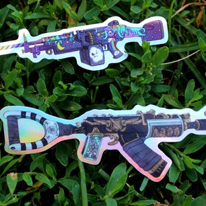 May include: Two holographic stickers of stylized assault rifles with intricate designs. The top sticker features a rainbow, stars, and a crescent moon. The bottom sticker features a black and white striped pattern, a bird, and a floral design.
