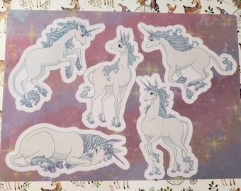 Op de afbeelding: Vijf witte eenhoorn stickers met blauwe manen en hoeven. De eenhoorns staan in verschillende poses, waaronder staand, rennend en liggend. De stickers zijn op een roze achtergrond met witte sterren.