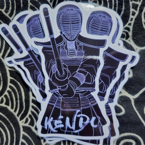 May include: A white sticker with a black outline of a person wearing a kendo uniform and holding a shinai. The text "KENDO" is written in white on the sticker.