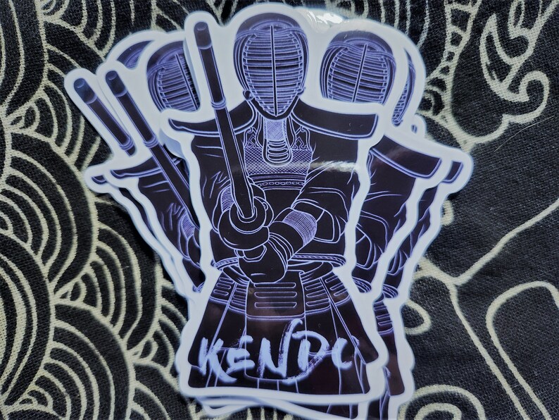 May include: A white sticker with a black outline of a person wearing a kendo uniform and holding a shinai. The text "KENDO" is written in white on the sticker.