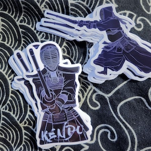 May include: A stack of white stickers with a black outline of a person wearing a kendo uniform and holding a sword. The sticker says "KENDO" in white lettering.