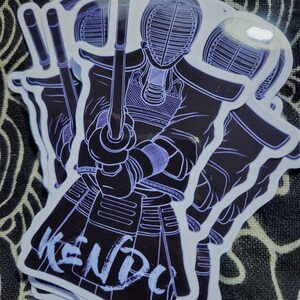 May include: A set of three black and white stickers featuring a kendo fighter in a traditional uniform holding a shinai. The stickers have a white outline and the word "KENDO" is written in white on the bottom sticker.
