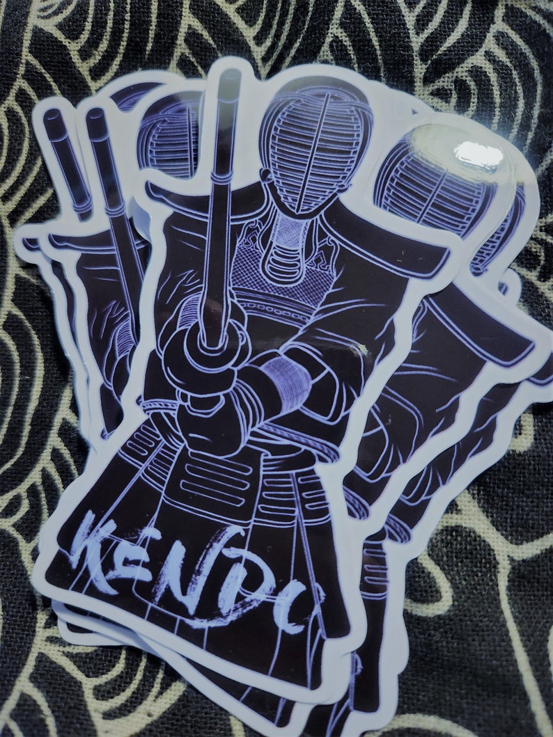 May include: A set of three black and white stickers featuring a kendo fighter in a traditional uniform holding a shinai. The stickers have a white outline and the word "KENDO" is written in white on the bottom sticker.
