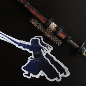 May include: Two blue and white stickers of a kendo practitioner in a fighting pose, holding a shinai sword. The stickers are on a black surface next to a decorative katana sword with a brown handle and a white and black scabbard.