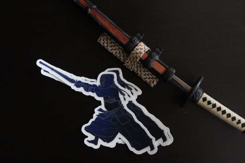 May include: Two blue and white stickers of a kendo practitioner in a fighting pose, holding a shinai sword. The stickers are on a black surface next to a decorative katana sword with a brown handle and a white and black scabbard.