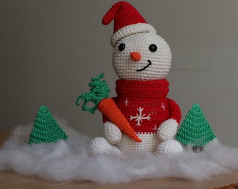 May include: A crocheted snowman wearing a red sweater with a snowflake design and a red Santa hat, holding a carrot. The snowman is sitting in a bed of white cotton, with two small green crocheted trees on either side.
