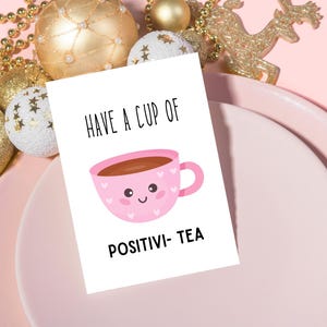 May include: A white card with the text "HAVE A CUP OF POSITIVI-TEA" and a pink teacup illustration. The teacup has a smiling face and heart details. The card is on a pink plate, surrounded by gold ornaments.