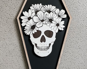 May include: A black and white skull with a floral wreath made of white flowers on a black background. The skull and flowers are framed in a wooden coffin-shaped frame.