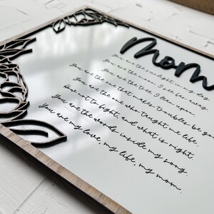 Op de afbeelding: Een wit houten bord met een zwart bloemenontwerp en de tekst "Mom" in grote zwarte letters. Het bord heeft een handgeschreven boodschap die luidt: "You are the sunlight in my day. You are the moon I see far away. You are the tree I lean upon. You are the one that makes troubles be gone. You are the one who taught me life... How not to fight, and what is right. You are the words inside my song. You are my love, my life, my mom."