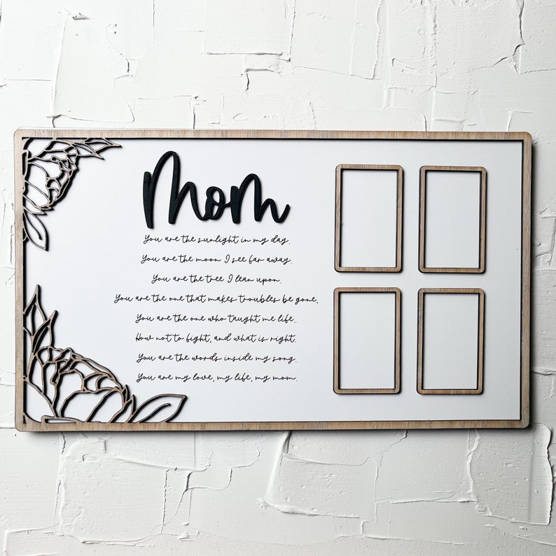 Op de afbeelding: Een wit houten bord met een zwarte uitsparing van het woord "mom" en een gedicht over moederschap. Het bord heeft vier rechthoekige ruimtes voor foto's.