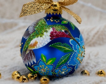 May include: A blue glass Christmas bauble with a hand-painted hummingbird and floral design. The bauble is decorated with gold glitter and has a gold bow and bells.