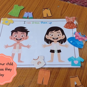 May include: A learning game for children featuring two paper dolls, a boy and a girl, with various clothing items to dress them up. The game includes a variety of clothing options, including shirts, pants, dresses, and accessories. The text on the game board reads "I can dress them up".