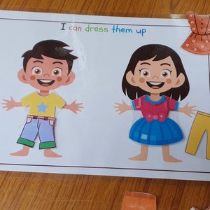 May include: A printable activity sheet with two paper dolls, a boy and a girl, and various clothing items to dress them up. The text on the sheet reads "I can dress them up".
