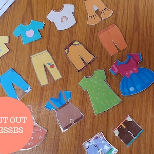 May include: A set of 14 colorful cut-out paper dress shapes, including a blue dress with a red bow, a green dress with white polka dots, and a yellow dress with a red apple. The paper shapes are on a wooden surface.