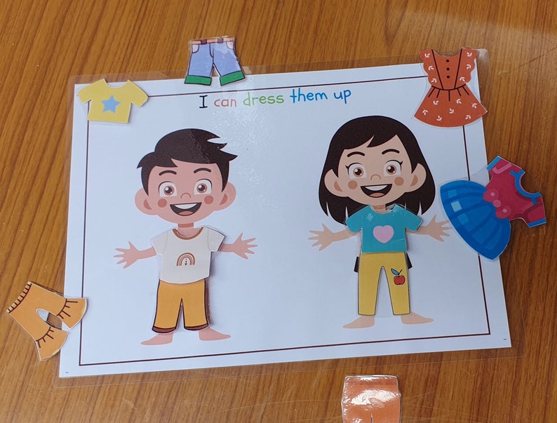 May include: A printable activity for kids featuring two cartoon characters, a boy and a girl, with various clothing items to dress them up. The text on the page reads "I can dress them up".
