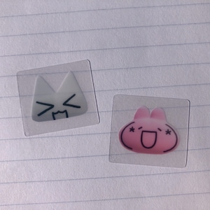 May include: Two clear stickers with cartoon faces. The first sticker is white with a cat face and the second sticker is pink with a bunny face.