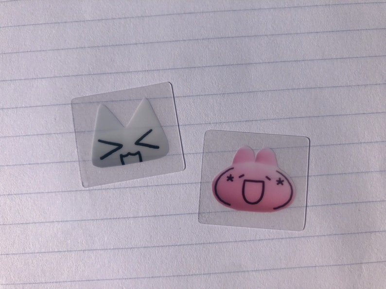 May include: Two clear stickers with cartoon faces. The first sticker is white with a cat face and the second sticker is pink with a bunny face.