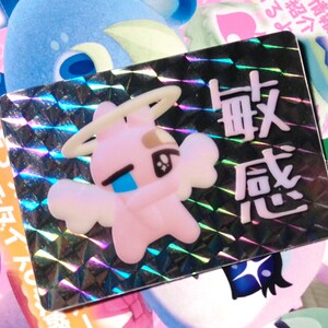 May include: A holographic sticker with a pink cartoon angel with a halo and wings. The sticker has Japanese text that reads "敏感" which translates to "sensitive".