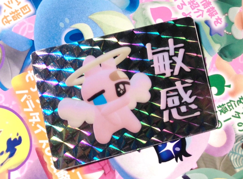 May include: A holographic sticker with a pink cartoon angel with a halo and wings. The sticker has Japanese text that reads "敏感" which translates to "sensitive".