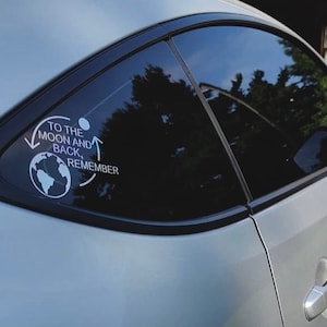 以下が含まれることがあります： 後ろの窓に「To the moon and back, remember」と書かれたステッカーが貼られた銀色の車。ステッカーには地球と三日月の小さな画像が描かれている。