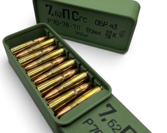May include: An olive green rectangular plastic storage box containing eight brass-colored cartridges arranged in a single row. The lid and side of the box feature black printed text including 7.62, P70-78-711, and other Cyrillic characters.