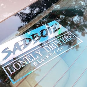 May include: A holographic sticker with the text "SADBOIZ" in a stylized font above the text "LONELY DRIVERS" in a white, rectangular frame. Japanese text is below the English text. The sticker is affixed to a car window.