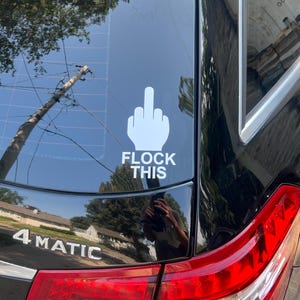 May include: A white vinyl decal applied to the rear window of a black vehicle. The decal features a graphic of a hand making a middle finger gesture positioned above the bold, white, sans-serif text that reads FLOCK THIS.