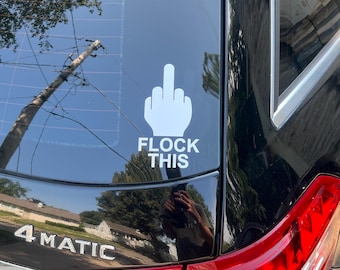 May include: A white vinyl decal applied to the rear window of a black vehicle. The decal features a graphic illustration of a hand making a middle finger gesture positioned above the bold, white, capitalized text that reads FLOCK THIS.