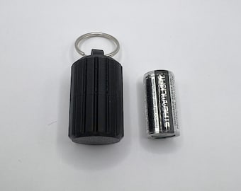 May include: Black cylindrical container with a key ring next to a silver Streamlight battery. The container has vertical grooves and a rounded base. The battery has black and silver text that reads "Streamlight".
