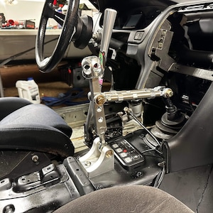 May include: A close-up of a custom-built shifter for a car. The shifter is made of metal and has a black knob with a skull design. The shifter is mounted to a black metal plate with a series of buttons.