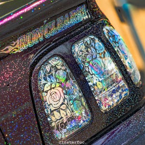 Könnte beinhalten: Nahaufnahme eines Fahrzeugfensters mit floralem Design. Die Fenster sind mit schillernden Rosen und Blättern verziert, vor einem dunklen, glitzernden Hintergrund. Die Texte "RUN CLEAR" und "clusterfuc." sind sichtbar.