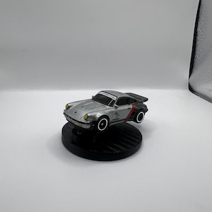 Op de afbeelding: Zilveren speelgoedauto met zwarte accenten, een rode streep en een achterspoiler. De auto heeft gele koplampen en staat op een zwarte, cirkelvormige basis. De wielen hebben witte velgen. Een miniatuur replica van een klassieke sportwagen.
