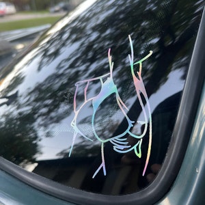 May include: A holographic sticker of a bird with its wings spread out. The sticker is on a car window.