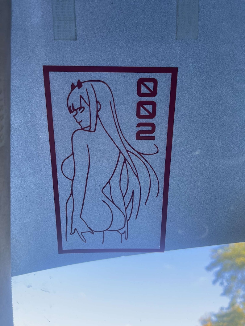 Peut inclure: Un dessin au trait noir et blanc d'un personnage d'anime avec de longs cheveux et un bandeau. Le personnage est debout, dos au spectateur. Le dessin est sur fond blanc avec le mot "002" en noir &agrave; droite du personnage.