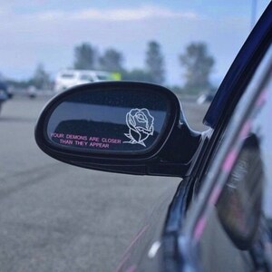 Peut inclure: Un r&eacute;troviseur de voiture noir avec un autocollant de rose blanche et le texte "Vos d&eacute;mons sont plus proches qu'ils n'en ont l'air" en lettres roses.