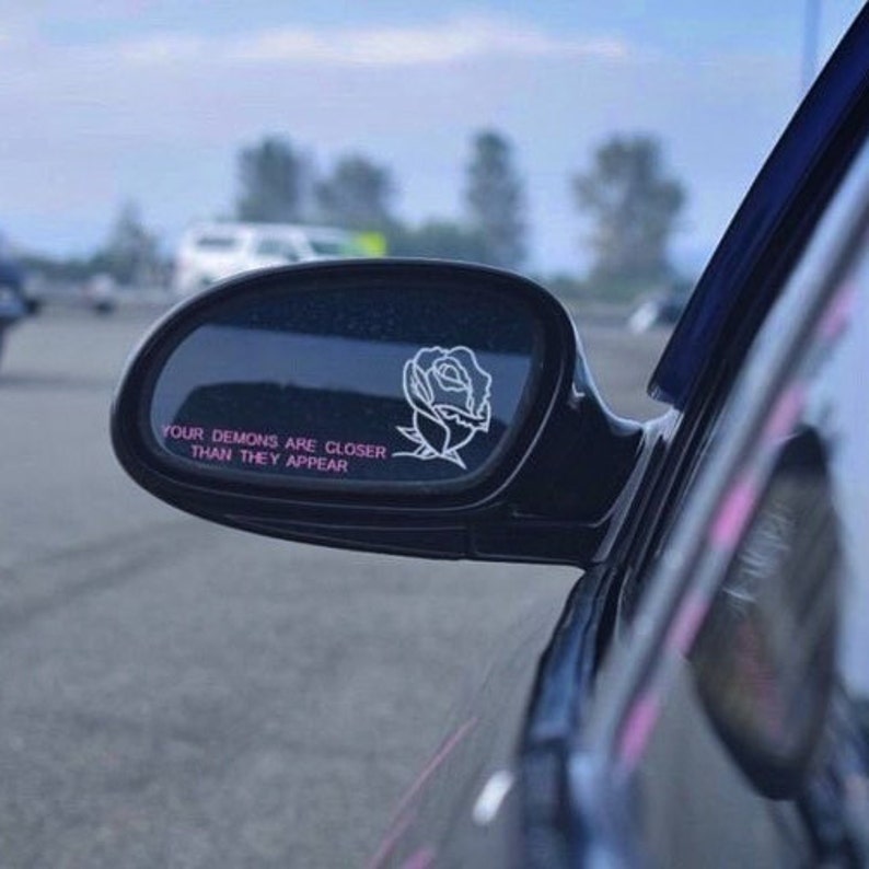 Peut inclure: Un r&eacute;troviseur de voiture noir avec un autocollant de rose blanche et le texte "Vos d&eacute;mons sont plus proches qu'ils n'en ont l'air" en lettres roses.