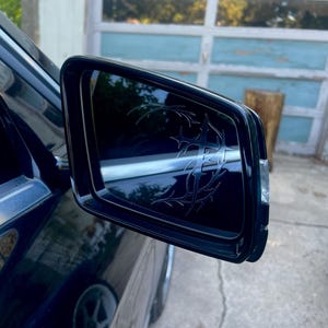 May include: Black side mirror with a decorative tribal design etched into the glass. The mirror is mounted on a dark blue car, reflecting the surrounding environment. The design is a series of interconnected lines and shapes.