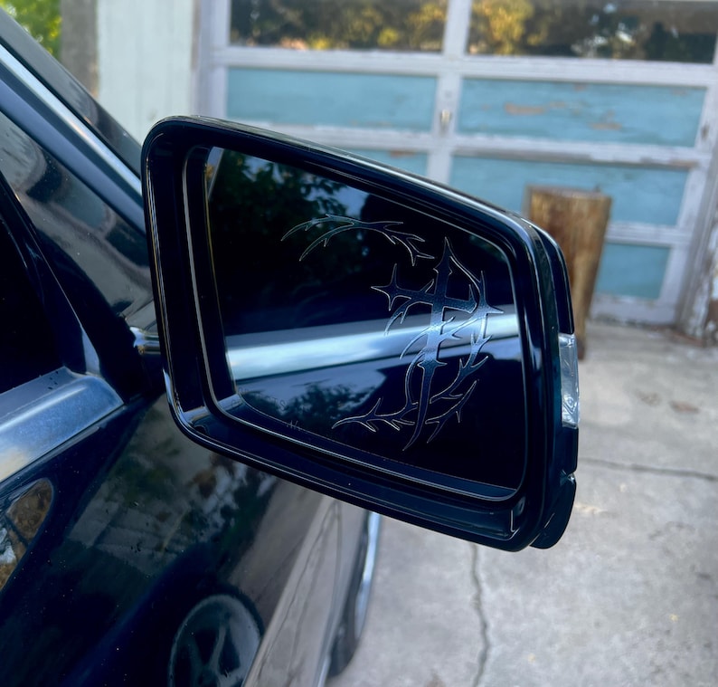 May include: Black side mirror with a decorative tribal design etched into the glass. The mirror is mounted on a dark blue car, reflecting the surrounding environment. The design is a series of interconnected lines and shapes.