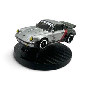 May include: A silver die-cast Porsche toy car featuring a black rear spoiler, yellow headlights, and black wheels with white rims. The windshield displays the text PORSCHE, and the side body includes black and red accents with small printed graphics.