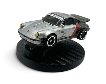 May include: A silver die-cast Porsche toy car featuring a black rear spoiler, yellow headlights, and black wheels with white rims. The windshield displays the text PORSCHE, and the side body includes black and red accents with small printed graphics.
