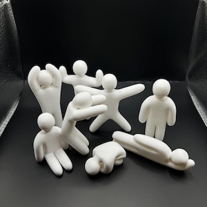 以下が含まれることがあります： 様々なポーズをとった7体の小さな白い3Dプリントフィギュアのセット。フィギュアは滑らかで丸みを帯びた頭部と手足をしており、立っているもの、座っているもの、横になっているものがあり、暗い背景に対して配置されています。