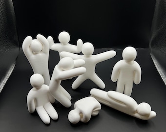 May include: A collection of seven small, white, 3D-printed figurines in various poses. The figures have smooth, rounded heads and limbs, with some standing, some sitting, and others lying down, all displayed against a dark, neutral background.
