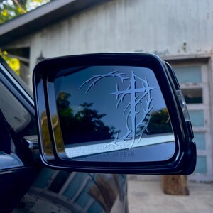 May include: Black side mirror with a decorative design featuring a cross and thorny vines. The mirror reflects a blurred outdoor scene. The text "OBJECTS IN MIRROR ARE CLOSER THAN THEY APPEAR" is visible.