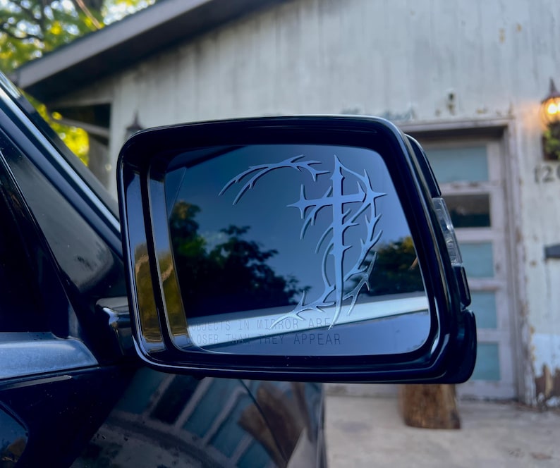 May include: Black side mirror with a decorative design featuring a cross and thorny vines. The mirror reflects a blurred outdoor scene. The text "OBJECTS IN MIRROR ARE CLOSER THAN THEY APPEAR" is visible.