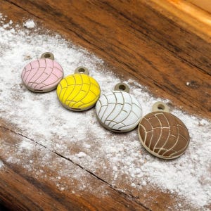 May include: Four colorful, round charms resembling concha pastries, each with a loop for hanging. The charms are pink, yellow, white, and brown, with a textured pattern. They are arranged on a wooden surface dusted with white powder.