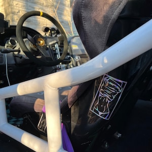 Puede incluir: Una jaula de seguridad blanca con una pegatina negra e iridiscente de un torso de mujer en el lateral. La pegatina tiene un diseño de un torso de mujer con los brazos cubriéndole la cara.