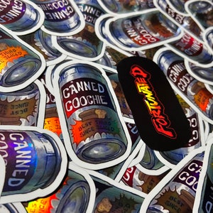May include: A close-up of several holographic stickers featuring a can of "Canned Goochie" with the text "Best Thing Since Sliced" and a holographic finish. The stickers are arranged in a pile, with some overlapping.